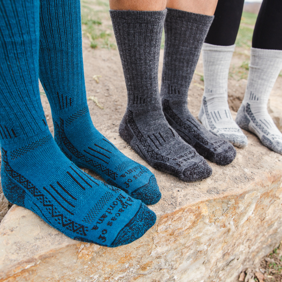 Three pairs of feet from the shins down stand on a rock, each wearing a pair of the Alpacas of Montana activewear outdoors athletic adventure hiking socks in blue, dark gray, and light gray (from left to right).