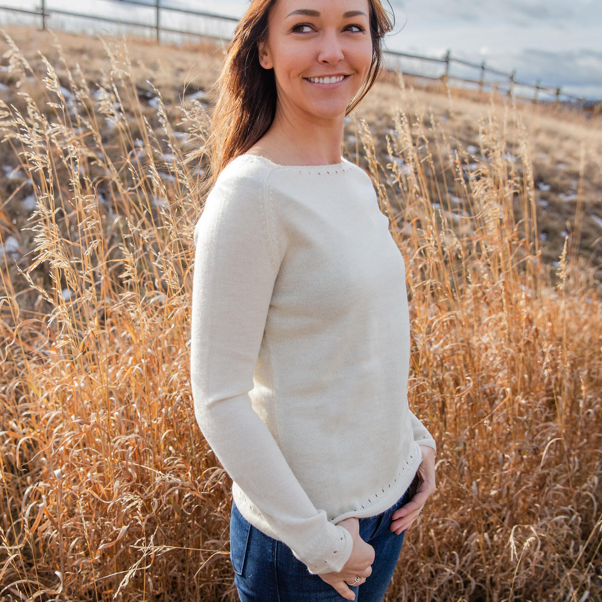 A woman standing in a field of golden grass looking off to the side of the frame. She is wearing blue jeans and the comfortable soft elegant natural white Alpacas of Montana Luxe Light sweater for women.