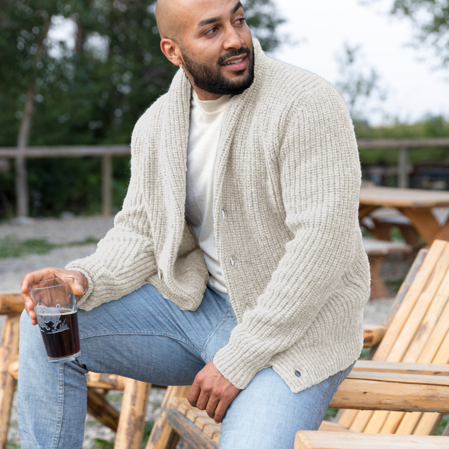 A bald man with a black beard looking off to the side while sitting on a wooden lounge chair holding a glass with a drink in it. He is wearing light blue jeans, a white shirt, and an Alpacas of Montana cozy comfortable soft moisture wicking formal casual stylish upcycled men&#39;s natural white button up chunky knit ribbed cardigan.
