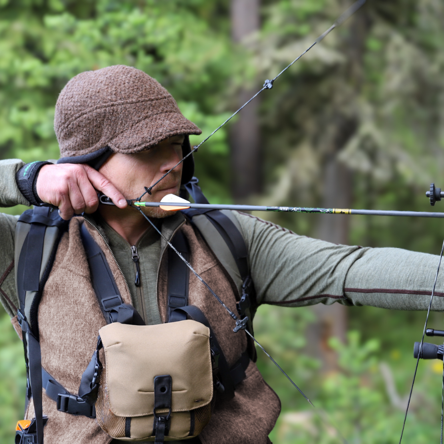Person aiming a bow and arrow outdoors while wearing a brown Extreme Warmth alpaca hat. The cozy textured alpaca material and structured ear coverage highlight the hat’s rugged, functional warmth in a forest setting.