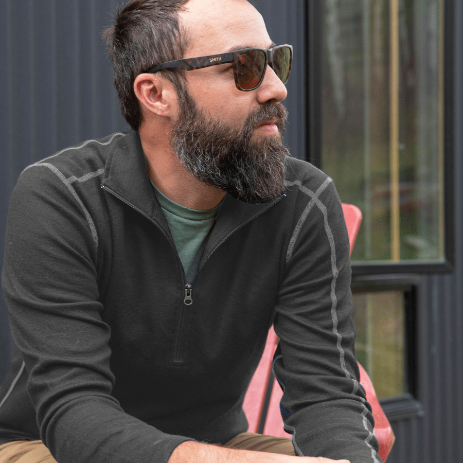A man with brown hair and a beard sitting on a chair and looking into the distance. He is wearing sunglasses and the Alpacas of Montana charcoal gray with light gray stitching warm soft cozy comfortable activewear outerwear athletic moisture wicking men&#39;s fashion stylish luxury quarter-zip alpaca wool top for outdoors camping hiking skiing hunting fishing running winter.
