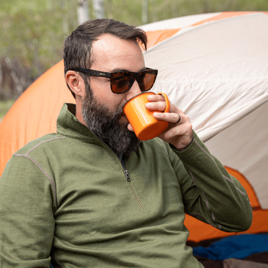 A man with brown hair and a beard sipping from an orange camping cup and sitting in front of a tent. He is wearing sunglasses and the Alpacas of Montana olive green warm soft cozy comfortable activewear outerwear athletic moisture wicking men&#39;s fashion stylish luxury quarter-zip alpaca wool top for outdoors camping hiking skiing hunting fishing running winter.
