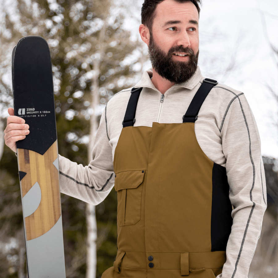 A man standing holding a pair of skis and looking into the distance. He is wearing a pair of light brown overall snowpants and a Alpacas of Montana oatmeal tan ivory with black stitching warm soft cozy comfortable activewear outerwear athletic moisture wicking men&#39;s fashion stylish luxury quarter-zip alpaca wool top for outdoors camping hiking skiing hunting fishing running winter.