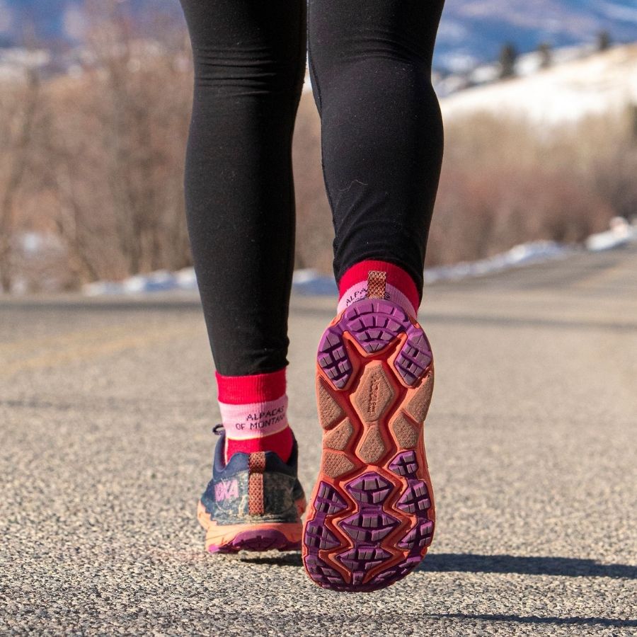 A knees-down photo of a person walking on a paved path in running shoes. They are wearing fuchsia, lime yellow, and light pink athletic moisture wicking activewear outerwear hiking quarter ankle socks.