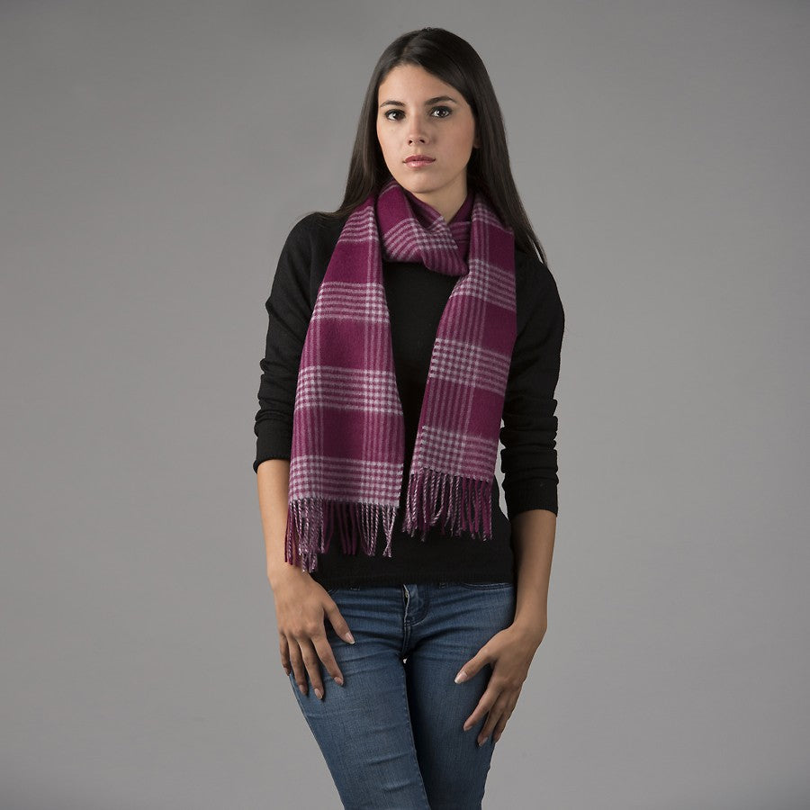 A photo of a woman with long black hair standing in front of a gray background. She is wearing blue jeans, a long sleeve black shirt, and an Alpacas of Montana soft stylish women&#39;s fashion comfortable cozy cute warm alpaca wool pink and plum plaid pattern scarf with tassels.