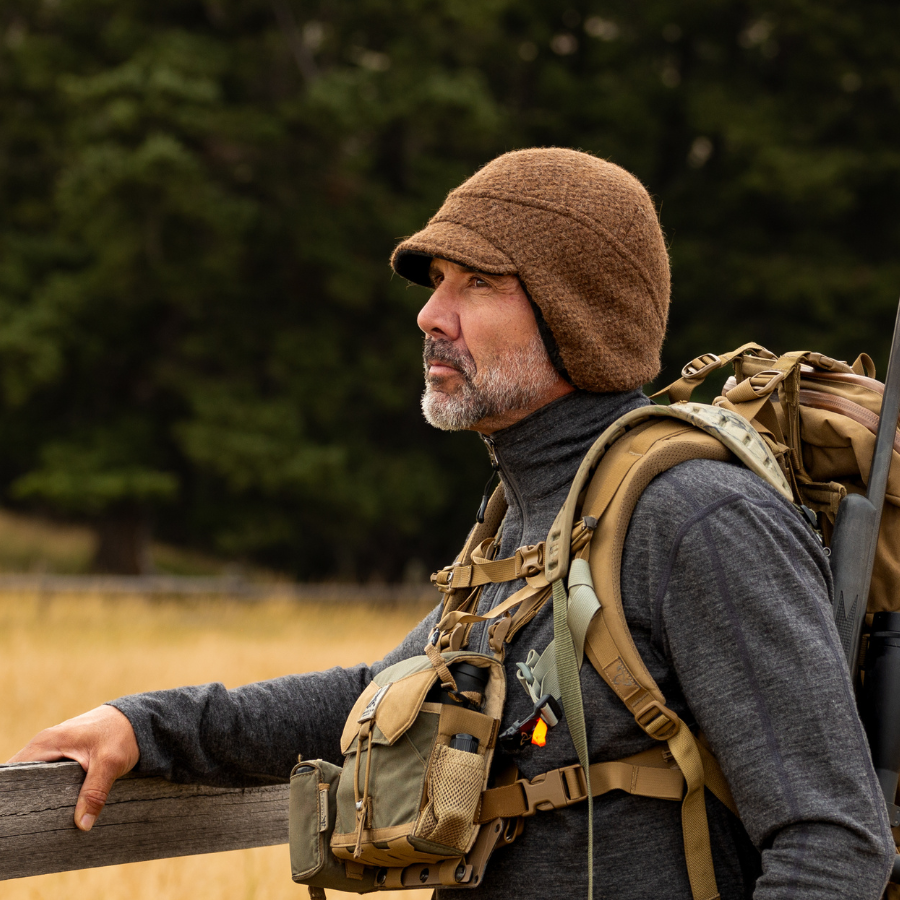 A man with a beard looking to the left wearing a backpack and brown alpaca hat with earflaps and a brim