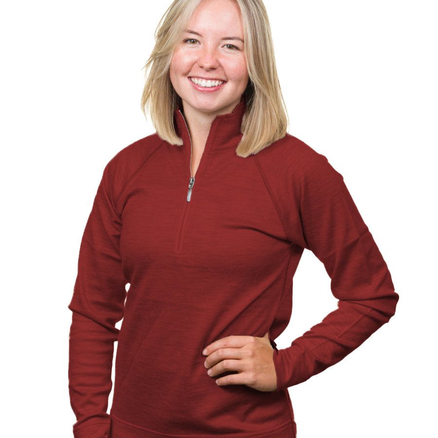 A waist-up photo of a smiling blonde woman standing in front of a white background with one hand on her hip. She is wearing a cranberry cherry red Alpacas of Montana warm soft cozy comfortable activewear outerwear athletic moisture wicking women&#39;s fashion stylish luxury quarter-zip alpaca wool top for outdoors camping hiking skiing hunting fishing running winter.