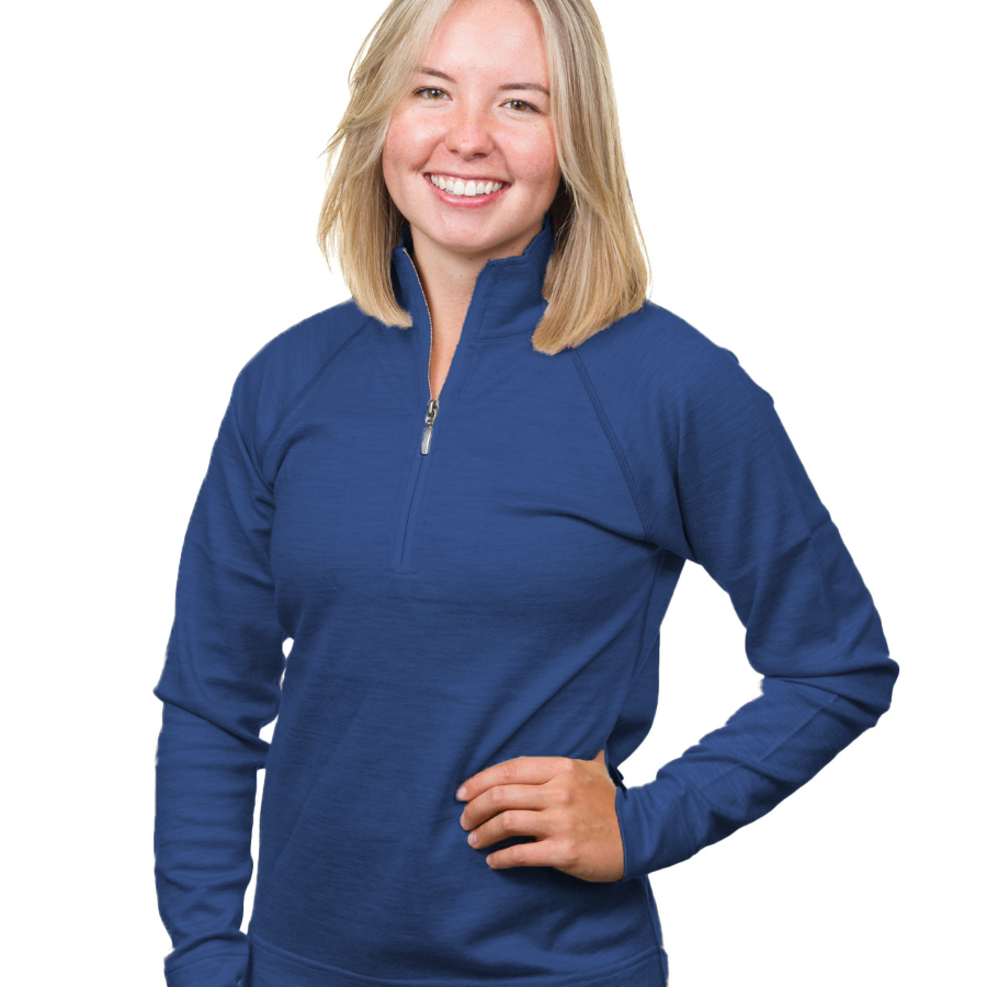 A waist-up photo of a smiling blonde woman standing in front of a white background with one hand on her hip. She is wearing a navy ocean blue Alpacas of Montana warm soft cozy comfortable activewear outerwear athletic moisture wicking women&#39;s fashion stylish luxury quarter-zip alpaca wool top for outdoors camping hiking skiing hunting fishing running winter.