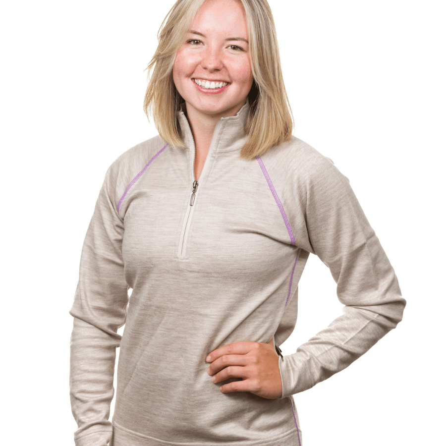 A waist-up photo of a smiling blonde woman standing in front of a white background with one hand on her hip. She is wearing a oatmeal tan ivory with purple stitching Alpacas of Montana warm soft cozy comfortable activewear outerwear athletic moisture wicking women&#39;s fashion stylish luxury quarter-zip alpaca wool top for outdoors camping hiking skiing hunting fishing running winter.
