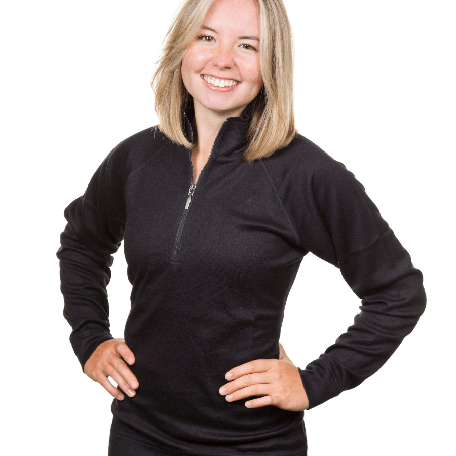 A waist-up photo of a smiling blonde woman standing in front of a white background with her hands on her hips. She is wearing a black Alpacas of Montana warm soft cozy comfortable activewear outerwear athletic moisture wicking women&#39;s fashion stylish luxury quarter-zip alpaca wool top for outdoors camping hiking skiing hunting fishing running winter.