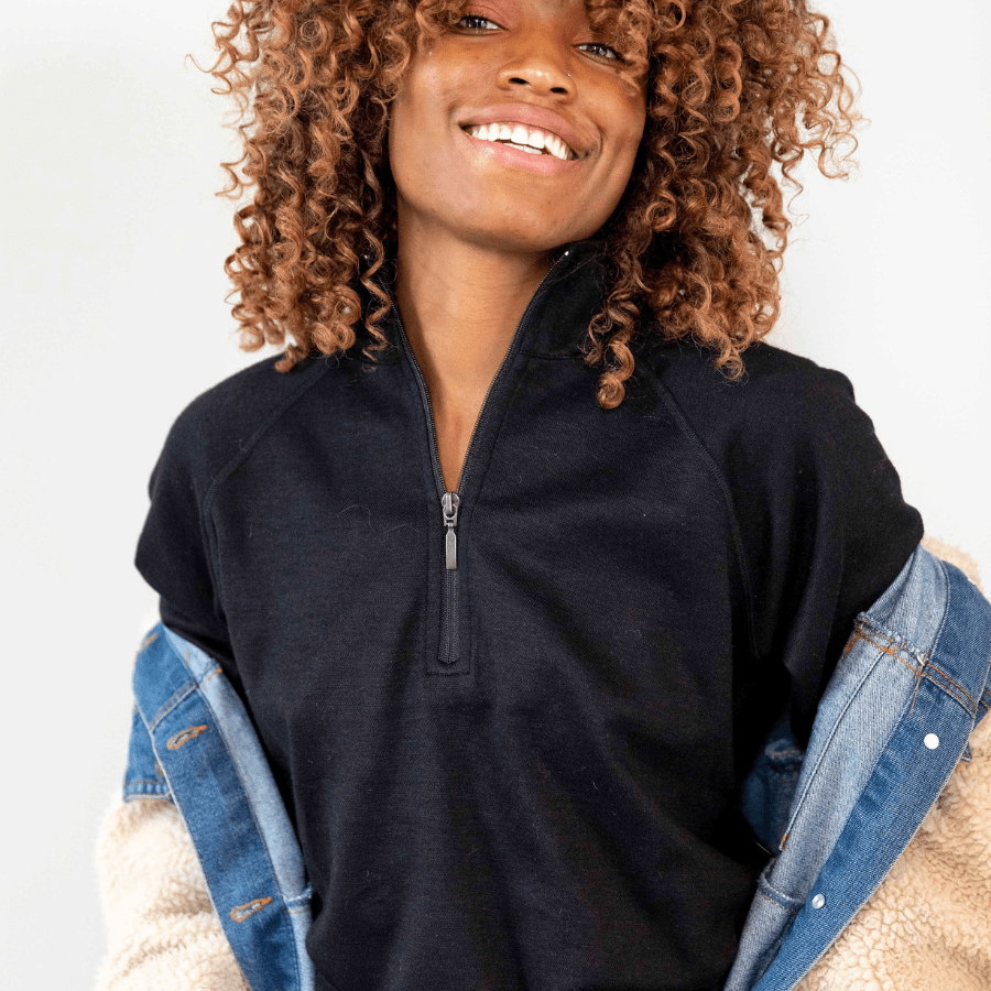 A smiling woman with curly brown hair in front of a white background. She is wearing a fleece and denim jacket and a black Alpacas of Montana warm soft cozy comfortable activewear outerwear athletic moisture wicking women&#39;s fashion stylish luxury quarter-zip alpaca wool top for outdoors camping hiking skiing hunting fishing running winter.