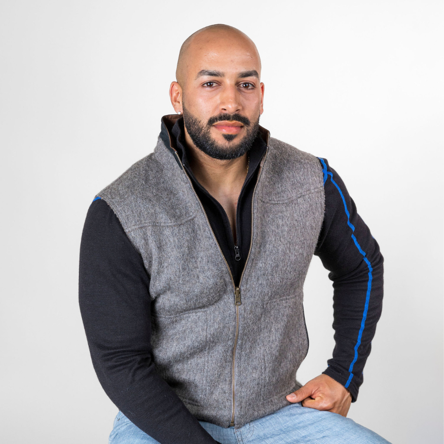 A bald man with a black beard sitting down against a white background and looking at the camera. He is wearing blue jeans, a black Alpacas of Montana men&#39;s base layer top, and a Alpacas of Montana camel brown and gray warm soft cozy comfortable neutral moisture wicking men&#39;s fashion stylish luxury zip manly reversible felted alpaca wool vest.