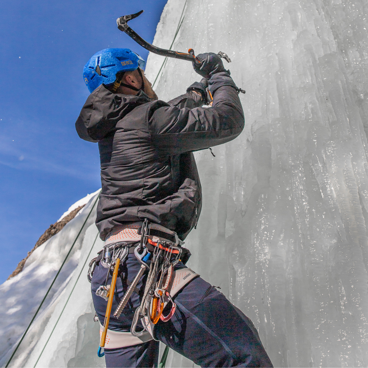 A photo looking from below a male ice climber wielding an ice pick. He is wearing a climbing harness belt with various clips, a pair of navy snowpants, black gloves, a bright blue helmet, and an Alpacas of Montana warm top layer insulated cozy comfortable thermal winter outerwear heavyweight alpaca wool lined graphite gray black granite peak expedition parka for skiing, snowboarding, ice fishing, hunting, camping, arctic, travel, outdoors
