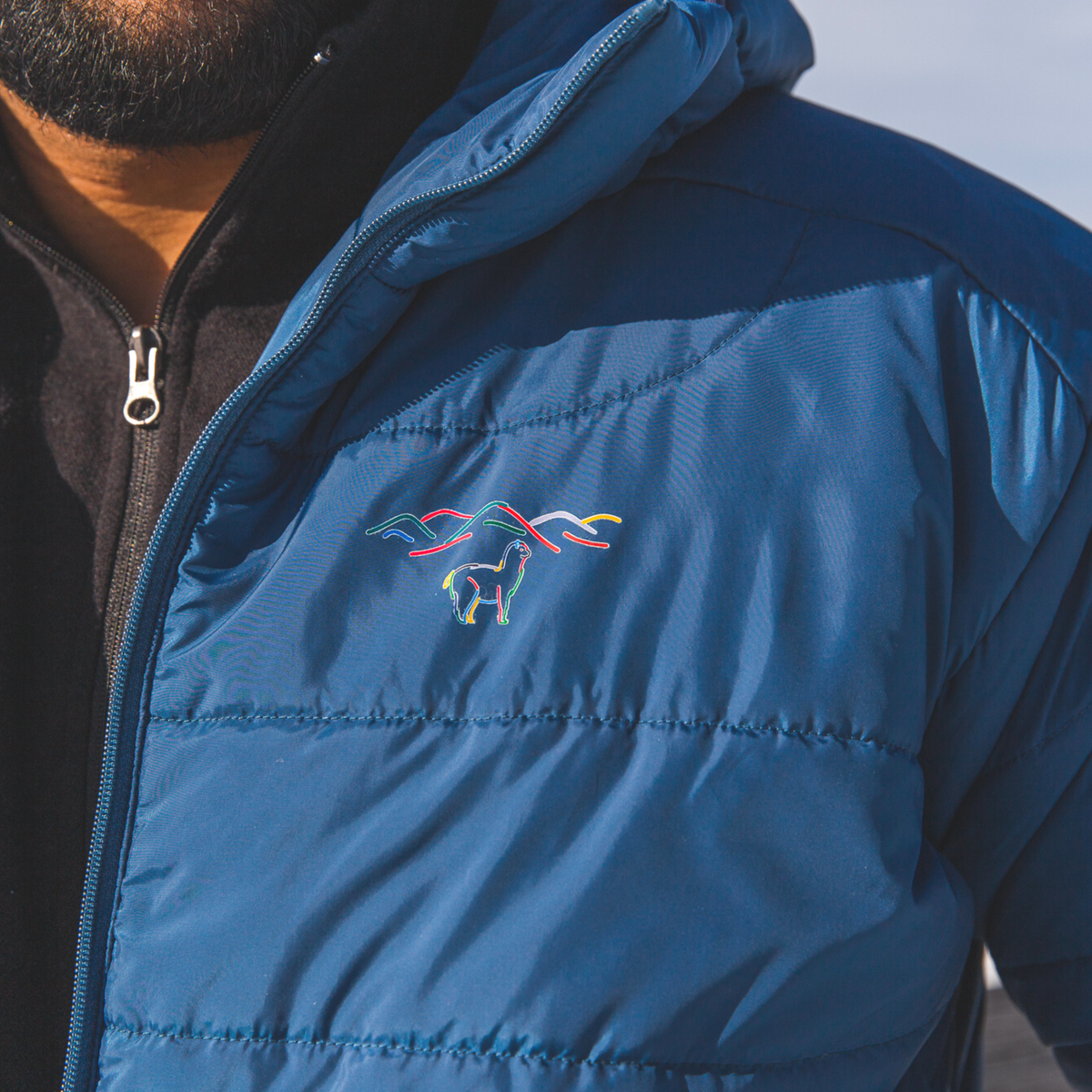 A close up of the Alpacas of Montana colorful logo on a cobalt blue Alpacas of Montana men&#39;s base layer top, and a Alpacas of Montana warm top layer insulated cozy comfortable thermal winter outerwear heavyweight alpaca wool lined men&#39;s mismi coat jacket parka for skiing, snowboarding, ice fishing, hunting, camping, arctic, travel, outdoors