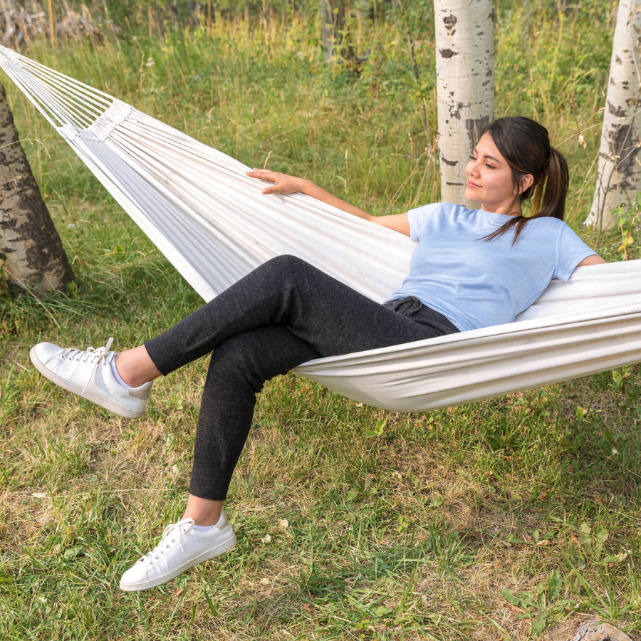 A smiling brown haired woman lounging in a hammock in a grassy forest wearing white sneakers, a light blue tee shirt, and A brown haired woman standing against a white background. She is wearing white sneakers, a white tank top, and charcoal gray Alpacas of Montana soft cozy comfortable lightweight lounge moisture wicking antimicrobial exercise women&#39;s 24/7 boyfriend joggers sweatpants made from alpaca wool for lounging, comfort, exercise, hiking, skiing
