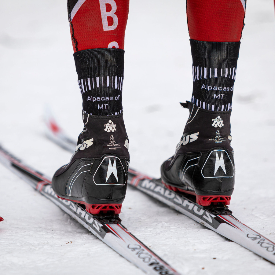 A close up photo of a person in boots on cross-country skis in the snow. They are wearing A photo of a person's lower legs standing against a white background wearing black, scarlet red, multi-gray, and white accent Alpacas of Montana soft cozy comfortable activewear outerwear athletic workout moisture wicking antimicrobial cushioned light compression engineered high-tech mid-crew hiking sock for walking, skiing, climbing, hunting, camping, fishing, exercise, biking