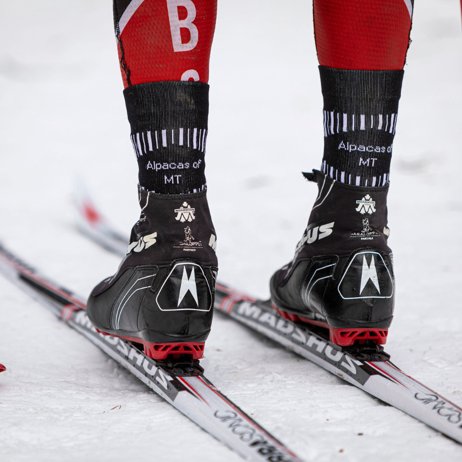 A close up photo of a person in boots on cross-country skis in the snow. They are wearing A photo of a person&#39;s lower legs standing against a white background wearing black, scarlet red, multi-gray, and white accent Alpacas of Montana soft cozy comfortable activewear outerwear athletic workout moisture wicking antimicrobial cushioned light compression engineered high-tech mid-crew hiking sock for walking, skiing, climbing, hunting, camping, fishing, exercise, biking
