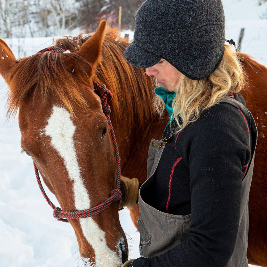 A blonde woman looking down at a horse standing next to her. She is wearing gray overalls, a black and red Alpacas of Montana women&#39;s mid-layer quarter zip top, and a dark gray Alpacas of Montana windstopper extreme warmth hat