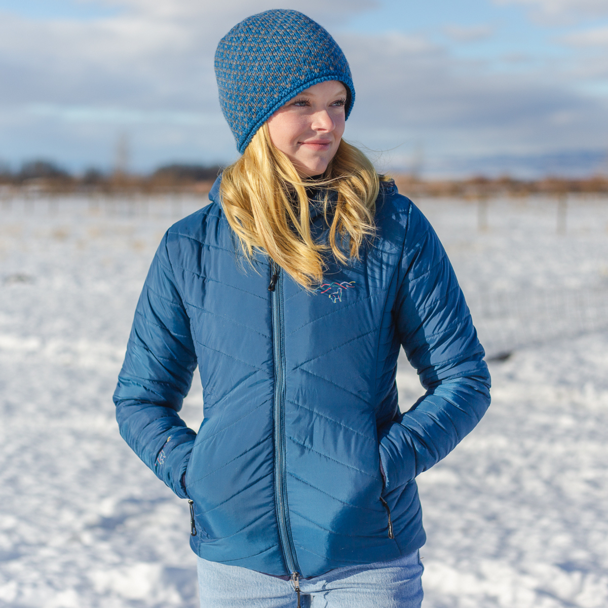 A blonde woman standing in a sunny winter scene. She is wearing a blue and gray Alpacas of Montana winter fleece beanie and a cobalt blue Alpacas of Montana men&#39;s base layer top, and a Alpacas of Montana warm top layer insulated cozy comfortable thermal winter outerwear heavyweight alpaca wool lined women&#39;s mismi coat jacket parka for skiing, snowboarding, ice fishing, hunting, camping, arctic, travel, outdoors