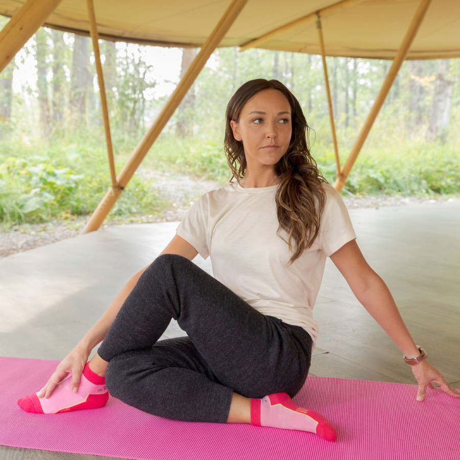 A brown haired woman sitting on a pink yoga mat underneath a canopy wearing a white tee shirt, Alpacas of Montana soft comfortable activewear charcoal gray 24/7 Boyfriend Joggers and fuchsia athletic Quarter Socks.