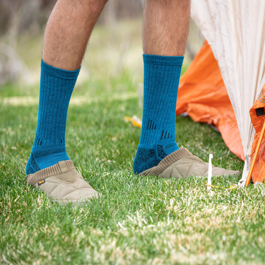 A photo from the shins down of feet standing near a tent wearing the blue activewear outdoors athletic adventure hiking socks and a pair of slippers.