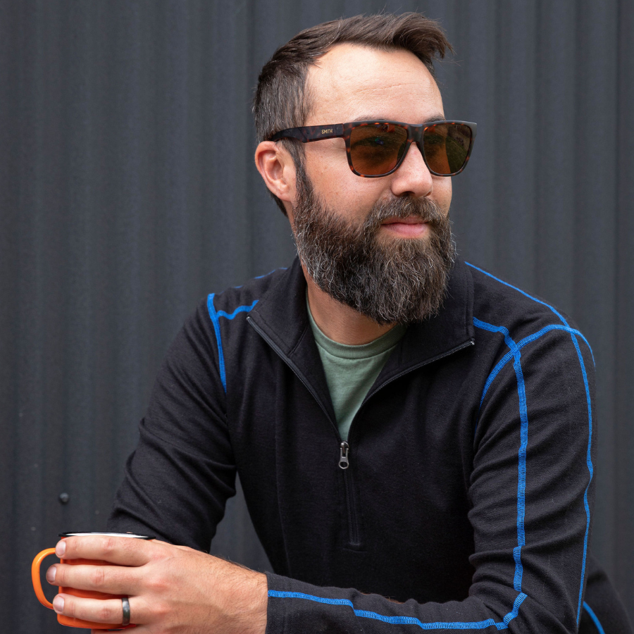 A man with brown hair and a beard looking off to the side and holding an orange camping mug. He is wearing a green tee shirt under a black with blue stitching warm soft cozy comfortable activewear outerwear athletic moisture wicking men&#39;s fashion stylish luxury mid-layer quarter-zip alpaca wool top for outdoors camping hiking skiing hunting fishing running winter.