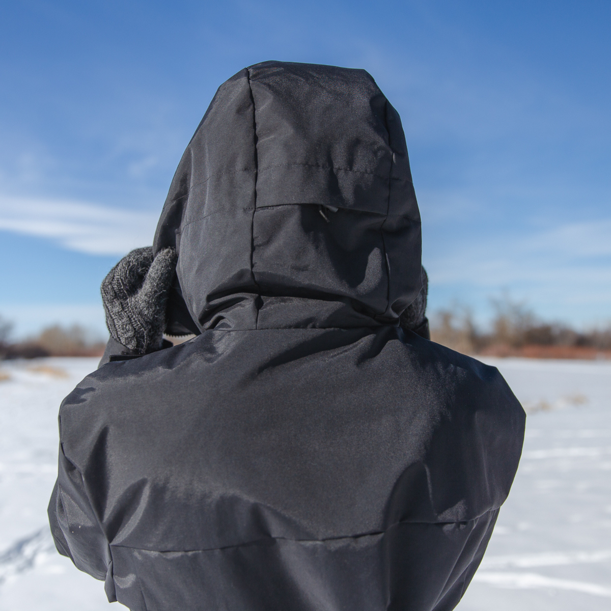 A close up photo of a person standing with their back to the camera in a sunny winter scene. They are wearing an Alpacas of Montana warm top layer insulated cozy comfortable thermal winter outerwear heavyweight alpaca wool lined graphite gray black granite peak expedition parka for skiing, snowboarding, ice fishing, hunting, camping, arctic, travel, outdoors