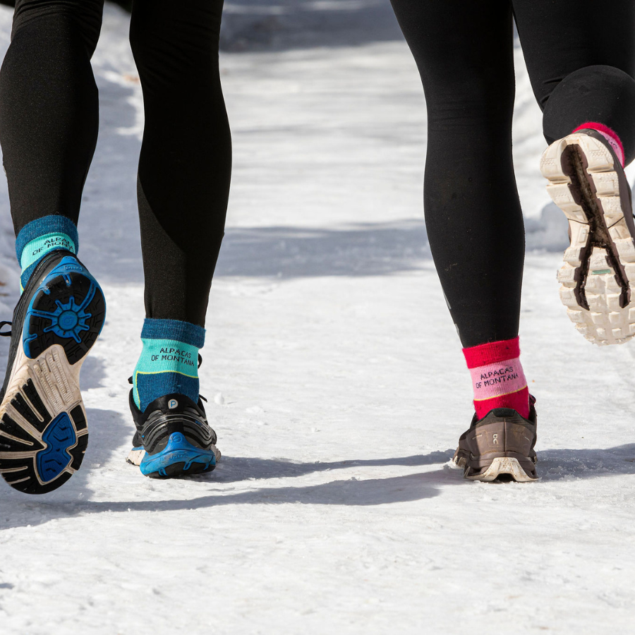 Two people running on a snowy path wearing tennis shoes. The person on the left is wearing teal, lime yellow, and navy blue Alpacas of Montana activewear athletic quarter socks. The person on the right is wearing fuchsia, lime yellow, and light pink Alpacas of Montana outerwear hiking quarter ankle socks.
