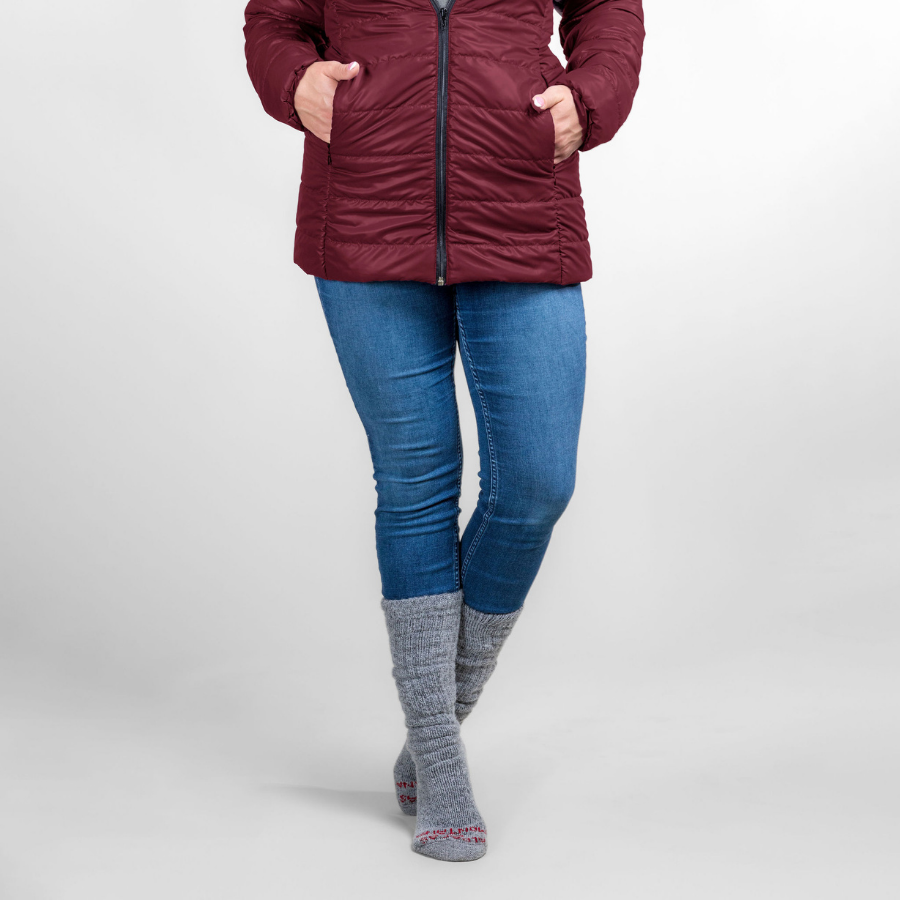 A waist-down photo of a woman standing in front of a white background wearing silver gray Alpacas of Montana arctic socks, blue jeans, and a cranberry cherry red Alpacas of Montana warm top layer insulated cozy comfortable thermal winter outerwear heavyweight alpaca wool lined electric peak parka jacket coat for skiing, snowboarding, ice fishing, hunting, camping, arctic, travel, outdoors