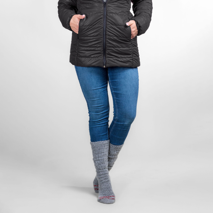 A waist-down photo of a woman against a white background wearing silver gray Alpacas of Montana arctic socks, blue jeans, and a black Alpacas of Montana warm top layer insulated cozy comfortable thermal winter outerwear heavyweight alpaca wool lined electric peak parka jacket coat for skiing, snowboarding, ice fishing, hunting, camping, arctic, travel, outdoors
