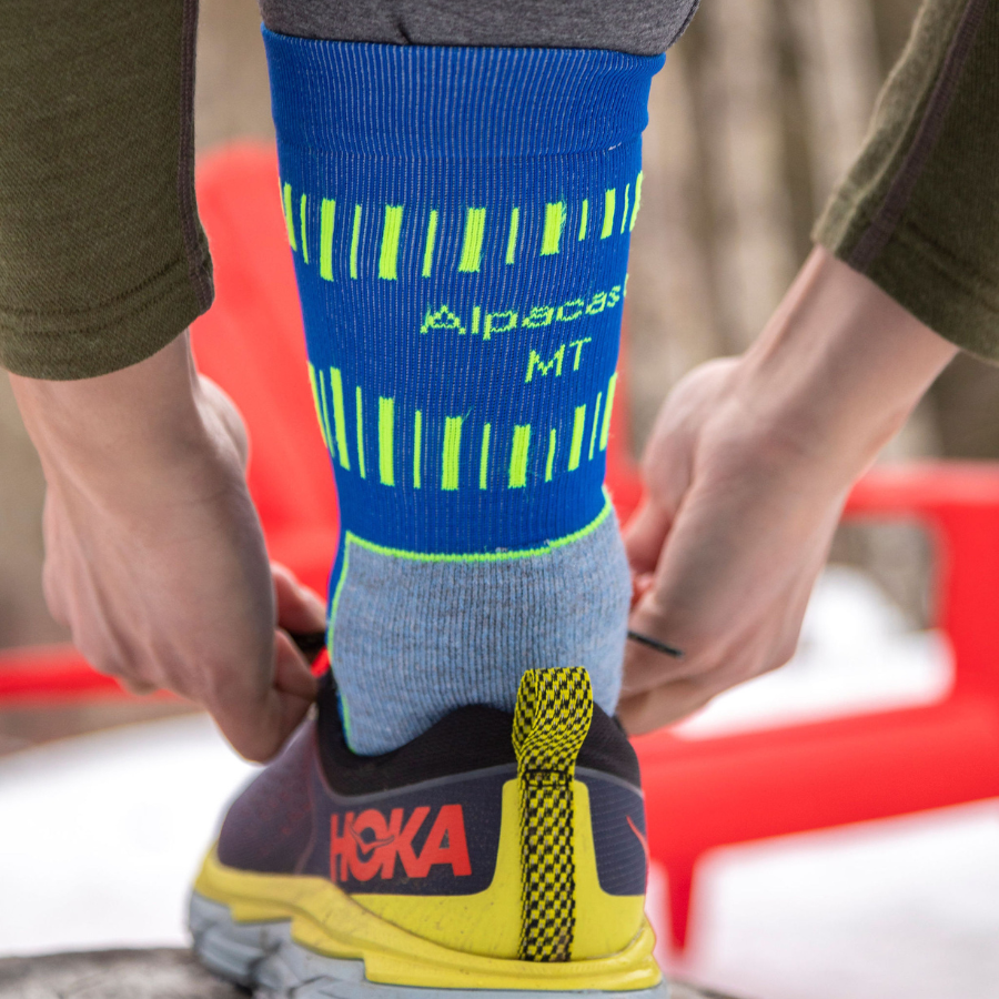 A close up photo of a person tying their running shoe wearing a cobalt royal blue, sky blue, and lime yellow Alpacas of Montana soft cozy comfortable activewear outerwear athletic workout moisture wicking antimicrobial cushioned light compression engineered high-tech mid-crew hiking sock for walking, skiing, climbing, hunting, camping, fishing, exercise, biking