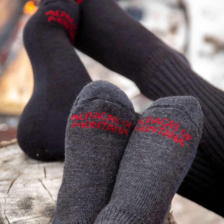 A close up photo of two pairs of feet on a stump. The closer pair of feet is wearing mid-calf soft comfortable cozy lounge breathable moisture wicking therapeutic diabetes loose fit dark gray socks, and the further pair is wearing mid-calf therapeutic socks in black.