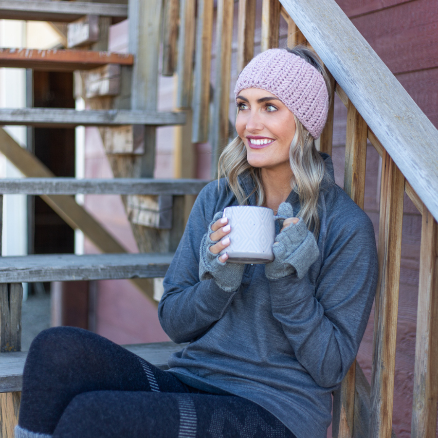 Blonde woman sitting on wooden steps looking to the side and holding a white mug. She is wearing dark gray pants, a medium gray long sleeve shirt, Alpacas of Montana lightweight flip mittens, and a light pink soft cute stylish fashion cozy comfortable warm winter headband handmade knit crochet in Montana from alpaca yarn