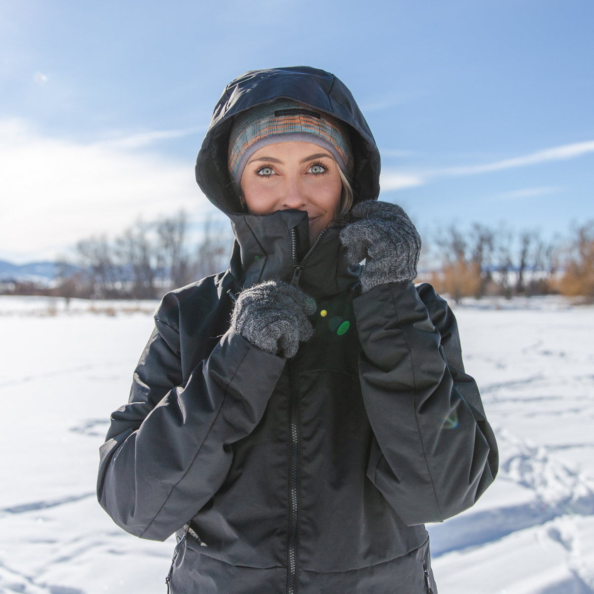 A photo of a woman standing in a sunny winter scene. She is wearing a teal, orange, and gray Alpacas of Montana backcountry beanie and an Alpacas of Montana warm top layer insulated cozy comfortable thermal winter outerwear heavyweight alpaca wool lined graphite gray black granite peak expedition parka for skiing, snowboarding, ice fishing, hunting, camping, arctic, travel, outdoors