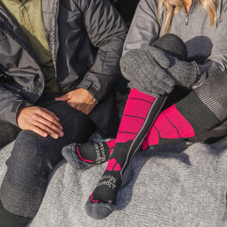 Two people sitting next to each other on an Alpacas of Montana gray boucle blanket. The person on the right is wearing a pair of pink, gray, and black Alpacas of Montana cozy comfortable soft warm thermal winter freezing temperatures antimicrobial moisture wicking alpaca wool ski and snowboard socks for snowshoeing, skiing, snowboarding, ice fishing, outdoors.