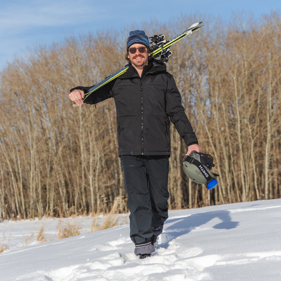 A man carrying a pair of skis over his shoulder and a ski helmet in his hand walking towards the camera through a snowy field with trees in the background. He is wearing black snowpants, an Alpacas of Montana multi-blue backcountry beanie, and an Alpacas of Montana warm top layer insulated cozy comfortable thermal winter outerwear heavyweight alpaca wool lined graphite gray black granite peak expedition parka for skiing, snowboarding, ice fishing, hunting, camping, arctic, travel, outdoors