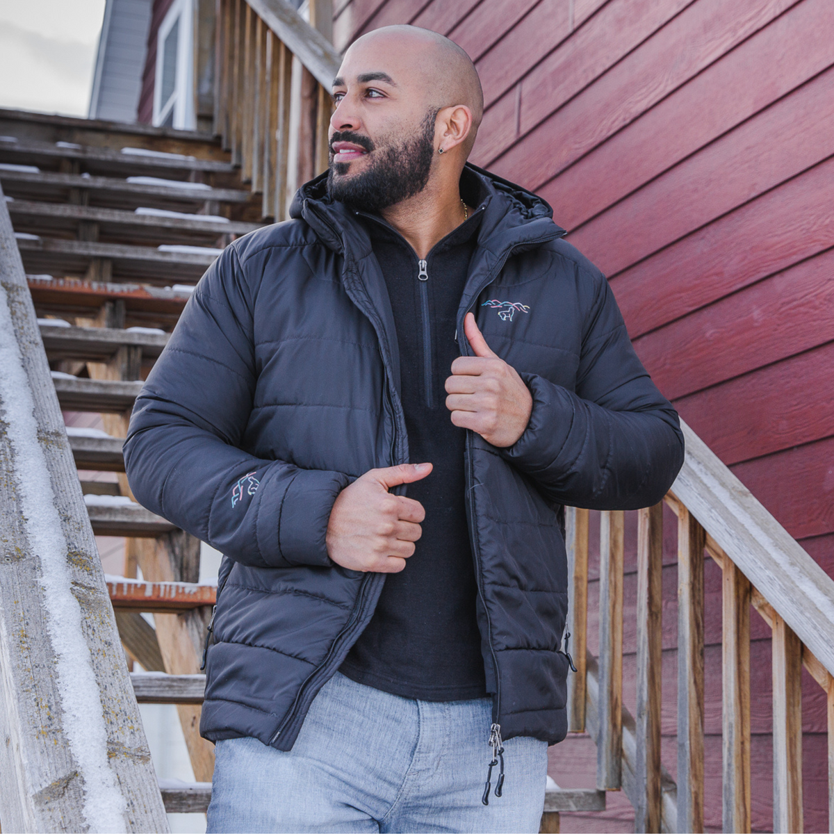 A photo of a bald man with a black beard standing on a set of wooden stairs looking into the distance. He is wearing blue jeans, a black Alpacas of Montana men&#39;s base layer top, and a Alpacas of Montana warm top layer insulated cozy comfortable thermal winter outerwear heavyweight alpaca wool lined men&#39;s mismi coat jacket parka for skiing, snowboarding, ice fishing, hunting, camping, arctic, travel, outdoors
