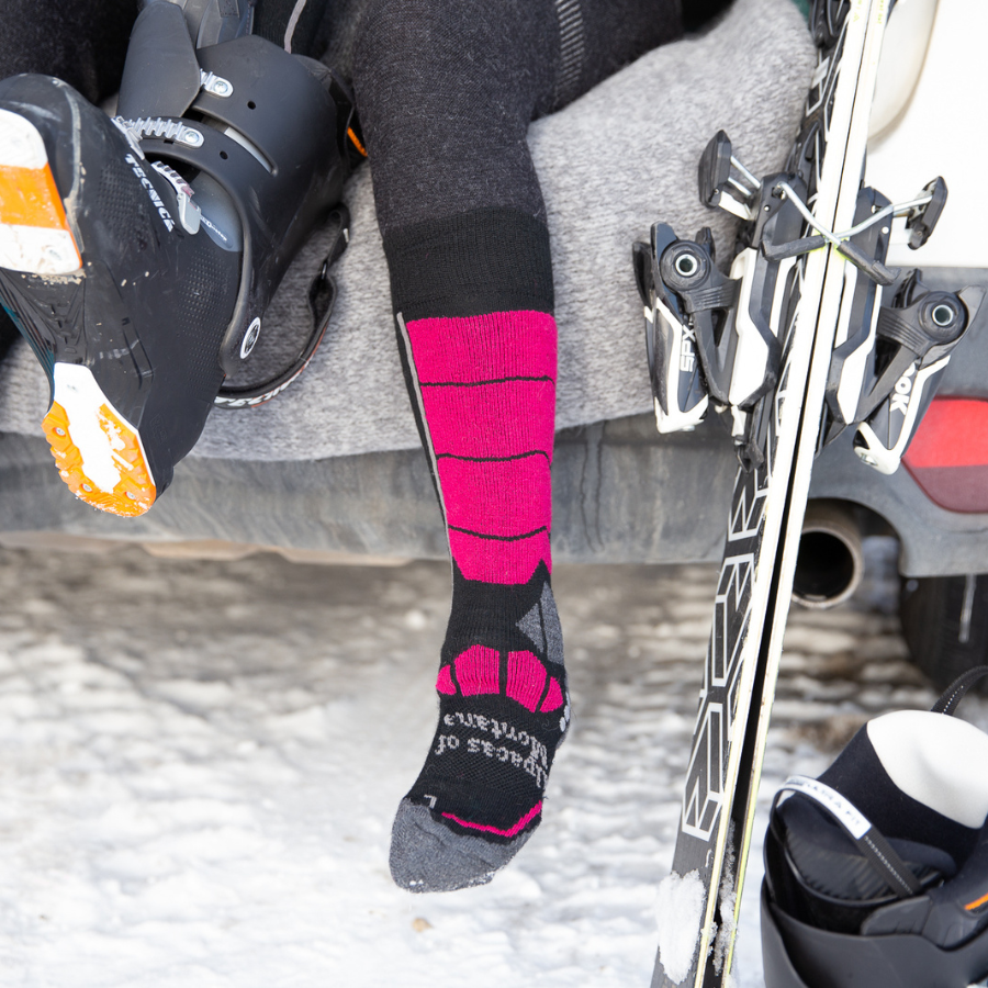 A person sitting in the back of a car on an Alpacas of Montana blanket holding a ski boot with skis resting nearby on the car. One foot is wearing a pink, gray, and black Alpacas of Montana cozy comfortable soft warm thermal winter freezing temperatures antimicrobial moisture wicking alpaca wool ski and snowboard socks for snowshoeing, skiing, snowboarding, ice fishing, outdoors.
