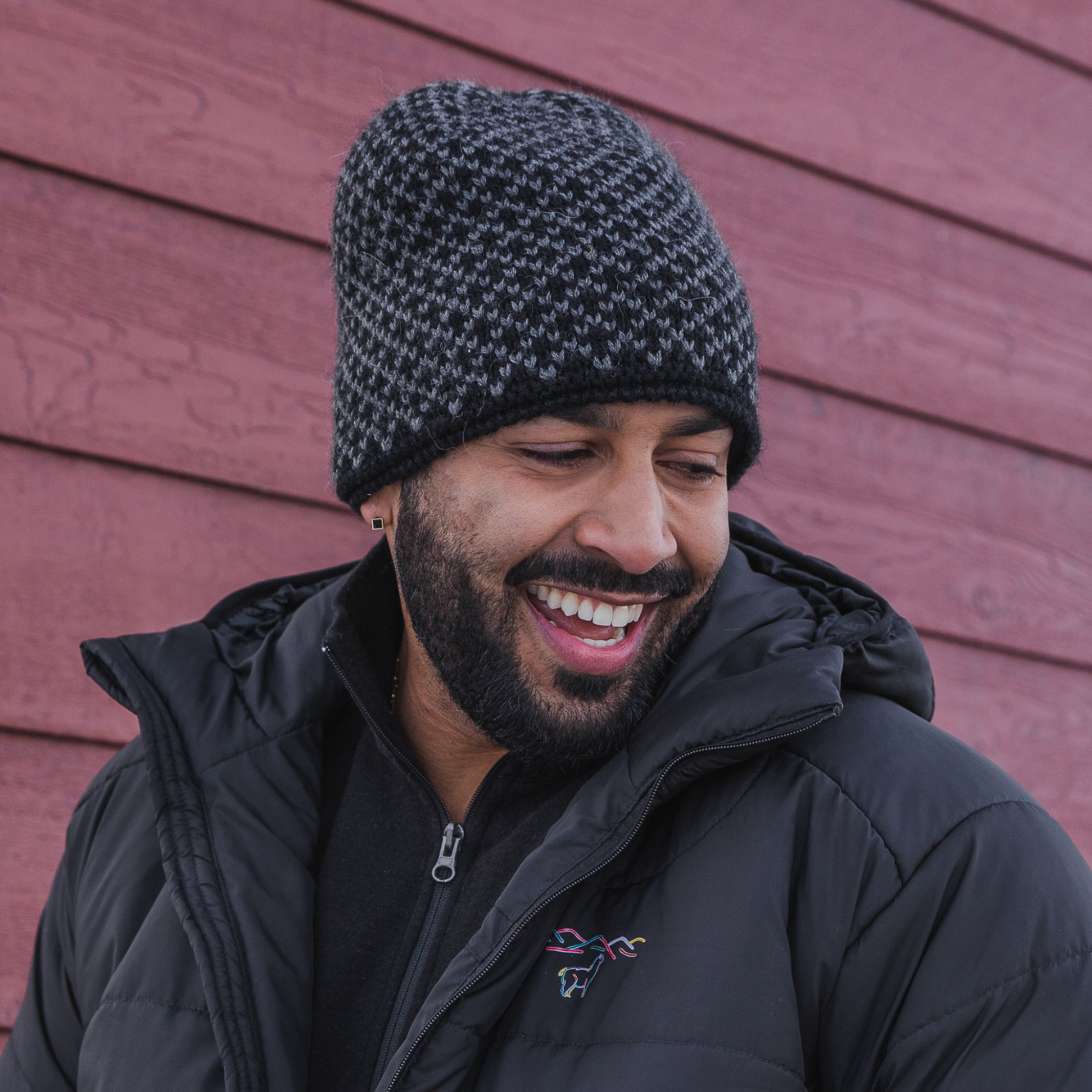 A smiling man with a black beard standing in front of a red barn and smiling at something off to the side of the frame. He is wearing a black Alpacas of Montana men's mid-layer quarter zip top, a black Alpacas of Montana granite peak expedition parka, and a black and gray Alpacas of Montana extremely warm cozy soft windproof comfortable antimicrobial moisture wicking thermal alpaca fleece wool winter fleece lined beanie hat for hiking, skiing, snowshoeing, sledding, outdoors.