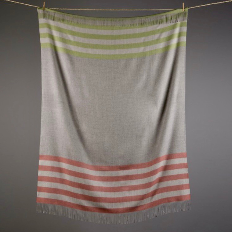 A gray background behind a clothesline with a hanging soft lightweight moisture wicking cozy comfortable lounge accessory interior design fun colorful Alpacas of Montana light gray, chartreuse green, and tangerine pink orange alpaca fleece wool throw blanket