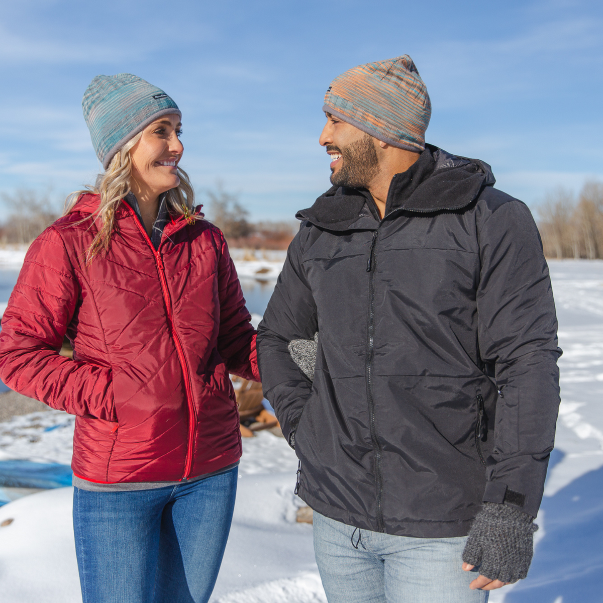 A woman and man smiling at each other and standing together in a sunny winter scene. The woman is wearing blue jeans, a cranberry red women&#39;s Electric Peak Parka, and a teal blue and gray soft comfortable warm winter hiking skiing outerwear Backcountry Beanie. The man is wearing handmade fingerless gloves, a graphite color Granite Peak Parka, and a teal, orange, and gray cozy moisture wicking Backcountry Beanie.