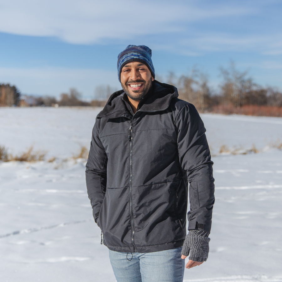 A photo of a smiling man with a black beard standing in a snowy field on a sunny winter day. He is wearing a multi-blue Alpacas of Montana backcountry beanie, Alpacas of Montana dark gray fingerless gloves, blue jeans, and an Alpacas of Montana warm top layer insulated cozy comfortable thermal winter outerwear heavyweight alpaca wool lined graphite gray black granite peak expedition parka for skiing, snowboarding, ice fishing, hunting, camping, arctic, travel, outdoors