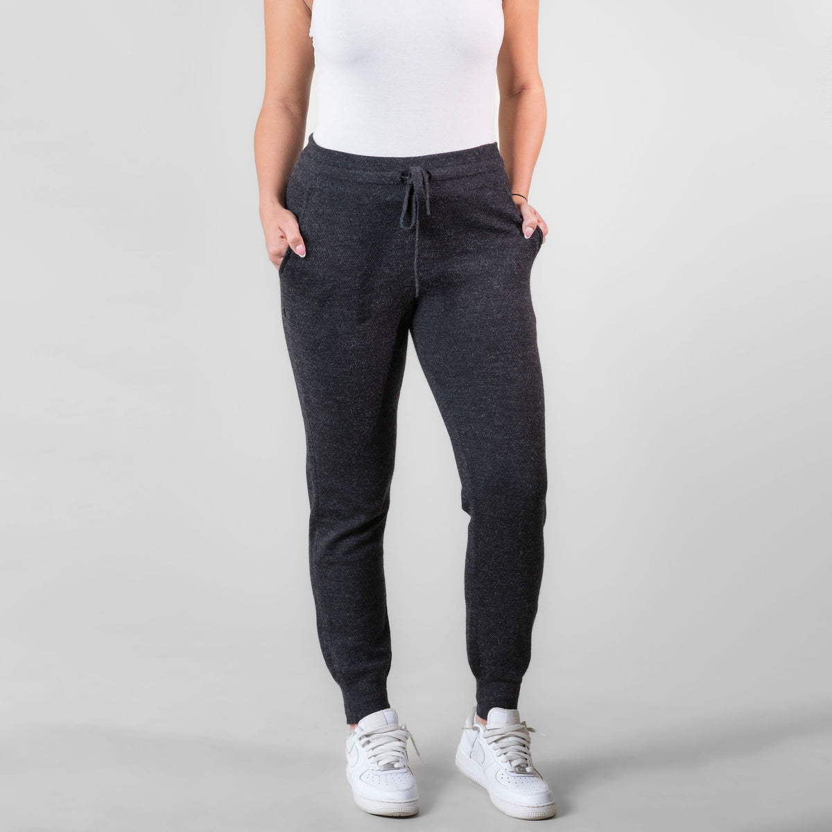 A bust-down photo of a woman standing against a white background. She is wearing white sneakers, a white tank top, and charcoal gray Alpacas of Montana soft cozy comfortable lightweight lounge moisture wicking antimicrobial exercise women&#39;s 24/7 boyfriend joggers sweatpants made from alpaca wool for lounging, comfort, exercise, hiking, skiing