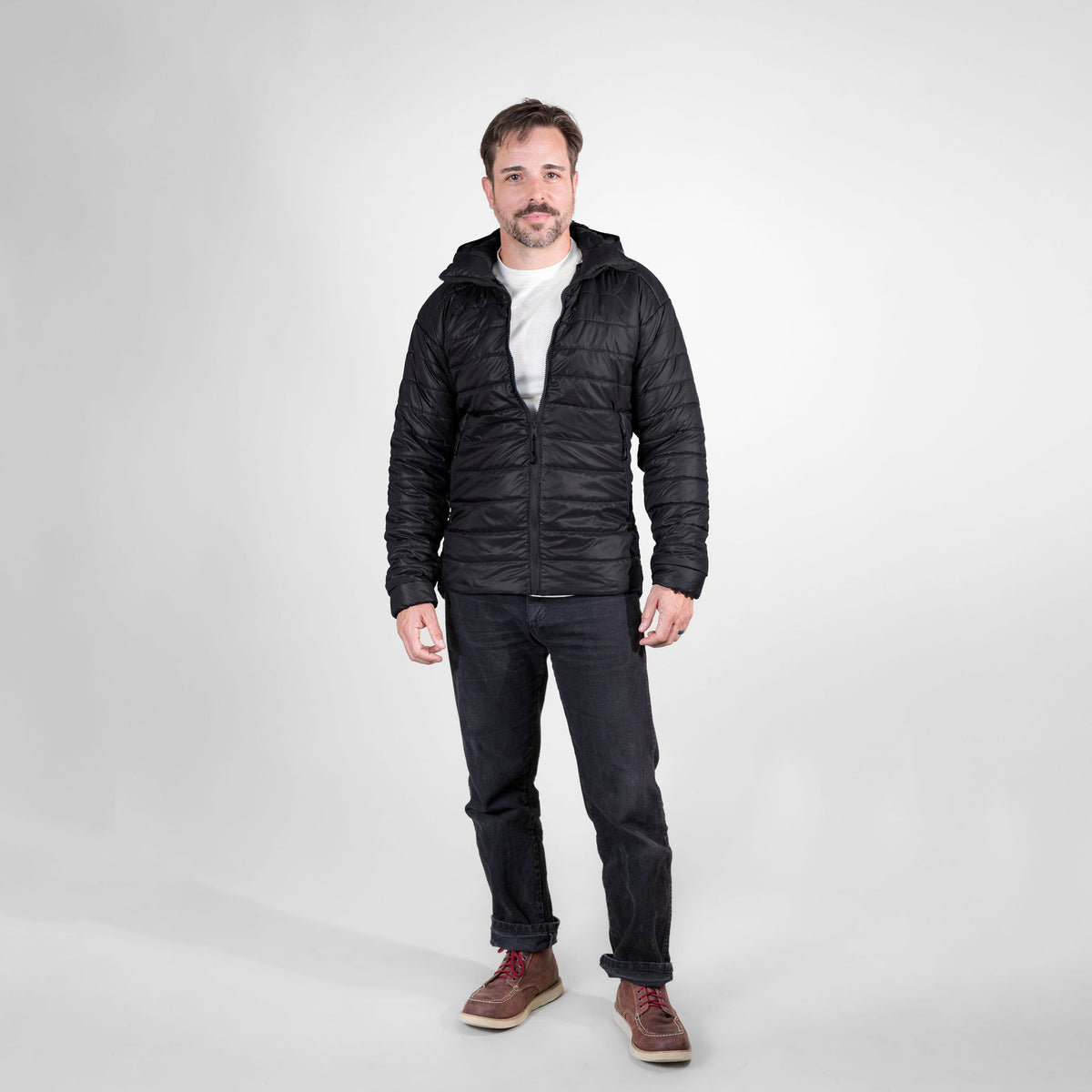 A man standing in front of a white background wearing brown boots, black jeans, a white tee shirt, and a black Alpacas of Montana men&#39;s base layer top, and a Alpacas of Montana warm top layer insulated cozy comfortable thermal winter outerwear heavyweight alpaca wool lined men&#39;s mismi coat jacket parka for skiing, snowboarding, ice fishing, hunting, camping, arctic, travel, outdoors