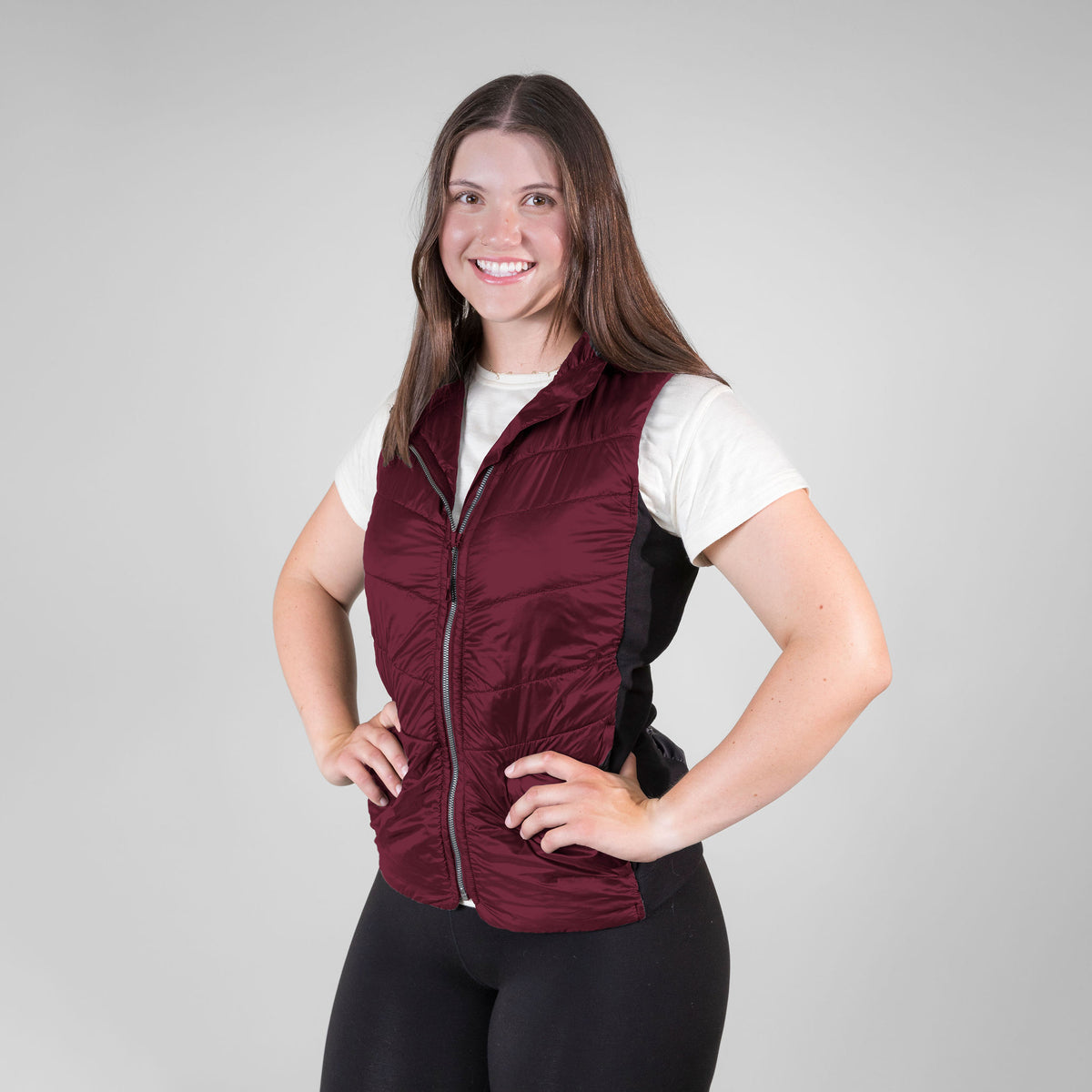 A smiling woman with brown hair standing with hands on hips in front of a white background. She is wearing black leggings, an Alpacas of Montana natural white women&#39;s performance tee, and a black Alpacas of Montana lightweight athletic activewear outerwear breathable moisture wicking antimicrobial soft comfortable exercise alpaca women&#39;s stretch lite vest for running sports exercise work out hiking climbing camping biking
