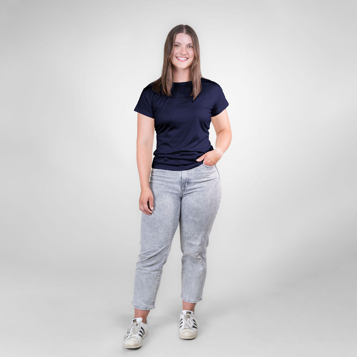 A full body photo of a smiling brown haired woman standing against a white background. She is wearing white sneakers, gray jeans, and a navy blue Alpacas of Montana lightweight athletic activewear outerwear breathable moisture wicking antimicrobial soft comfortable exercise short sleeve alpaca women&#39;s performance tee for running sports exercise work out hiking climbing camping skiing biking