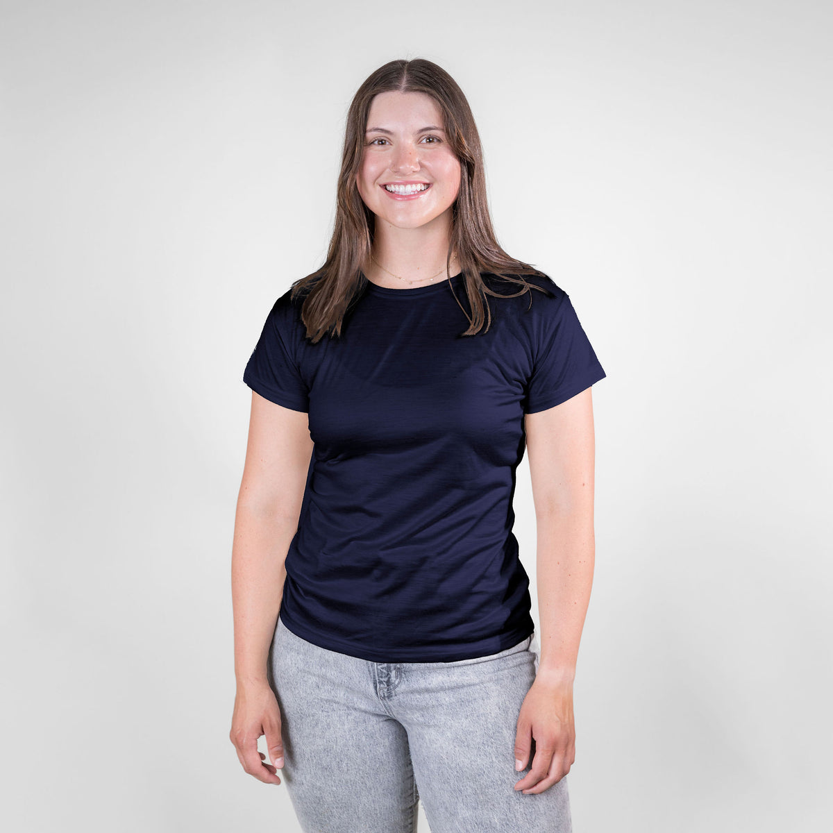 A smiling brown haired woman standing against a white background. She is wearing gray jeans and a navy blue Alpacas of Montana lightweight athletic activewear outerwear breathable moisture wicking antimicrobial soft comfortable exercise short sleeve alpaca women&#39;s performance tee for running sports exercise work out hiking climbing camping skiing biking