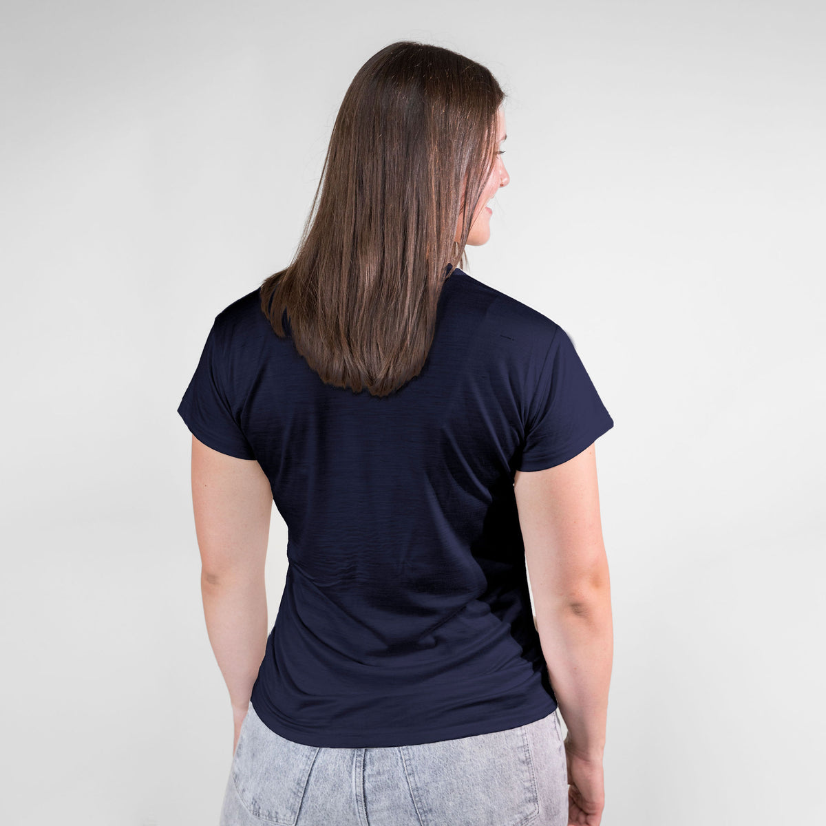 A brown haired woman facing away from the camera against a white background. She is wearing gray jeans and a navy blue Alpacas of Montana lightweight athletic activewear outerwear breathable moisture wicking antimicrobial soft comfortable exercise short sleeve alpaca women&#39;s performance tee for running sports exercise work out hiking climbing camping biking