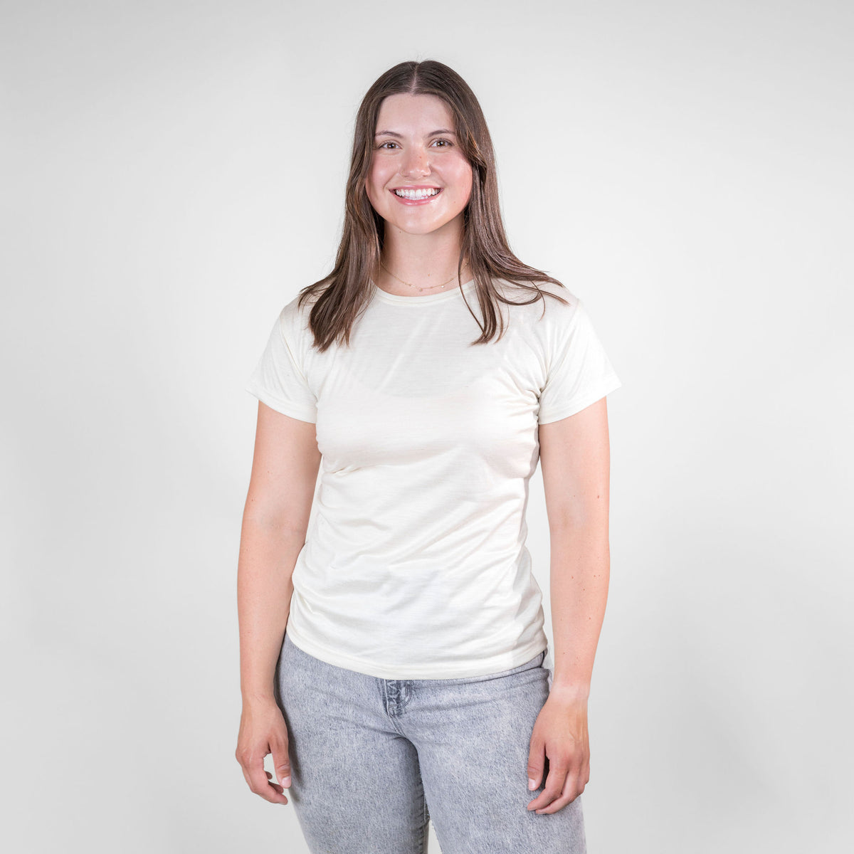 A smiling brown haired woman standing against a white background. She is wearing gray jeans and a natural white Alpacas of Montana lightweight athletic activewear outerwear breathable moisture wicking antimicrobial soft comfortable exercise short sleeve alpaca women&#39;s performance tee for running sports exercise work out hiking climbing camping biking