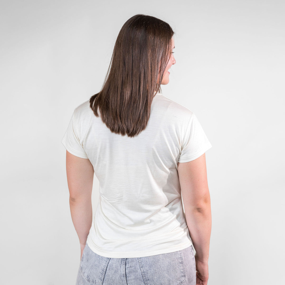 A brown haired woman facing away from the camera against a white background. She is wearing gray jeans and a natural white Alpacas of Montana lightweight athletic activewear outerwear breathable moisture wicking antimicrobial soft comfortable exercise short sleeve alpaca women&#39;s performance tee for running sports exercise work out hiking climbing camping biking